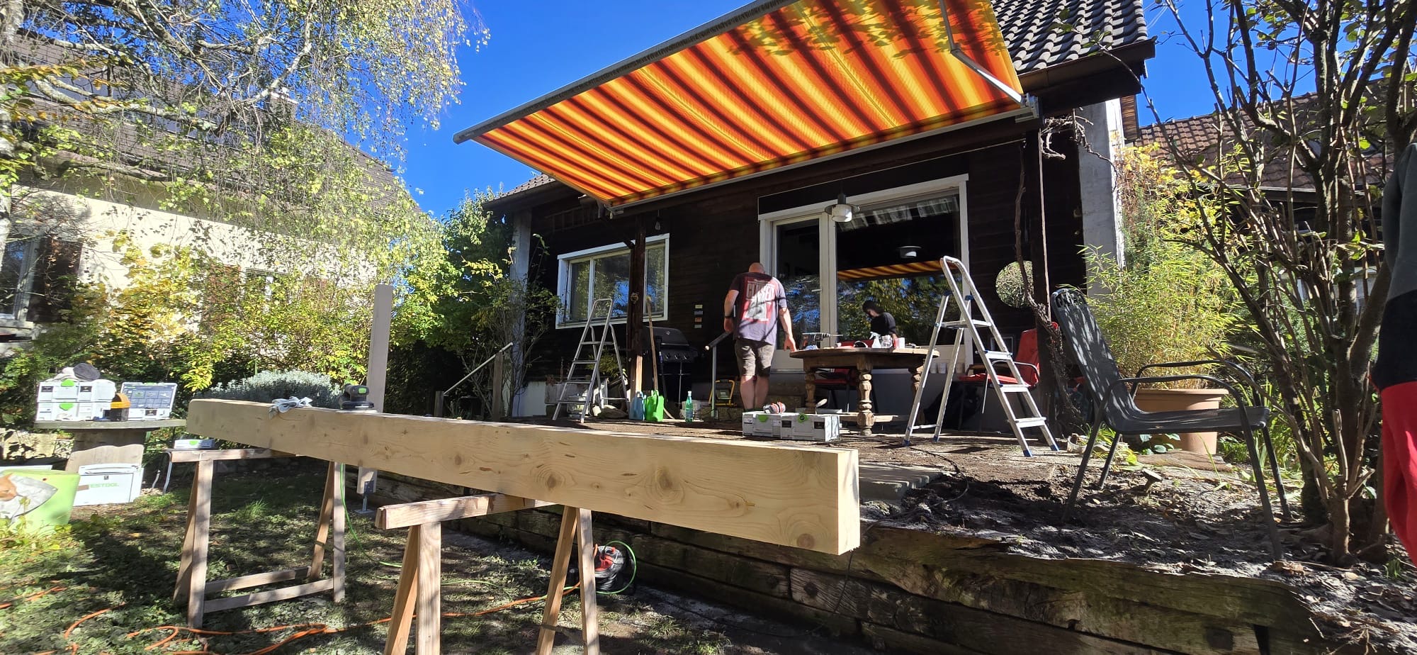 Unterkonstruktion einer Holzterrasse vor einem dunklen Holzhaus mit Markise